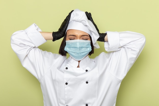 Free photo a front view young female cook in white cook suit and cap wearing gloves and sterile mask having severe headache on the green wall lady work food color