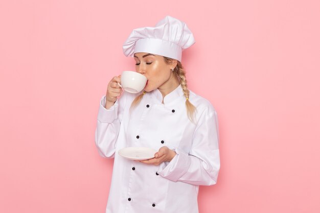 Front view young female cook in white cook suit drinking hot coffee on the pink space cook  