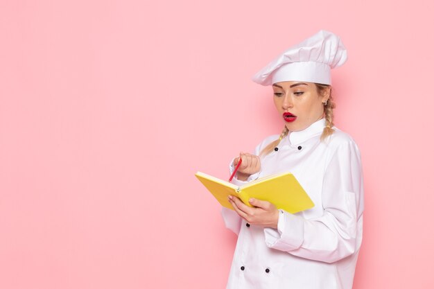 Front view young female cook in white cook suit holding yellow copybook and pencil on the pink space cook  food