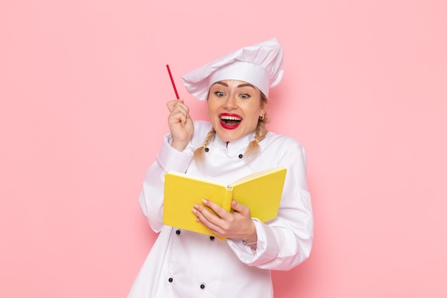 Front view young female cook in white cook suit holding yellow copybook and pencil on the pink space cook 