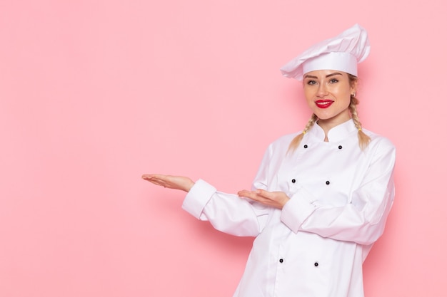 Front view young female cook in white cook suit slightly smiling and posing on the pink space cook  