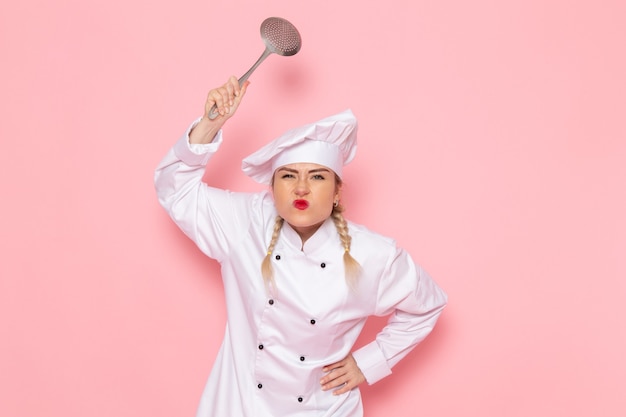 Free photo front view young female cook in white cook suit threatening with silver spoon on the pink space cook cuisine job work  photo