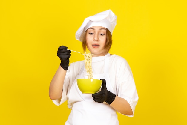 Free photo a front view young female cook in white cook suit and white cap in black gloves holding green plate mixing up spaghetti on the yellow