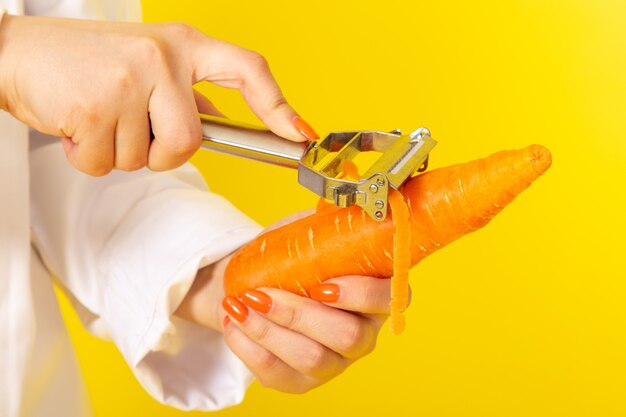 A front view young female cook in white cook suit and white cap holding and cleaning orange carrot on the yellow