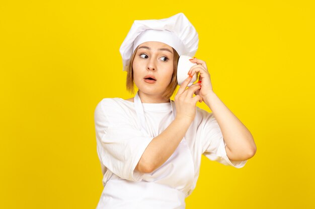 A front view young female cook in white cook suit and white cap holding white cup listening to cup on the yellow