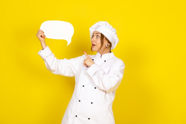 A front view young female cook in white cook suit and white cap holding white sign on the yellow