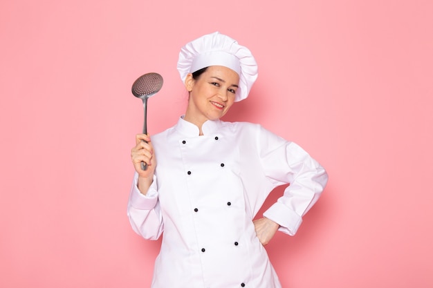 A front view young female cook in white cook suit white cap posing holding big silver spoon smiling