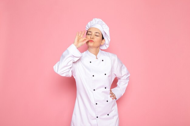 A front view young female cook in white cook suit white cap smiling posing delicious sign happy