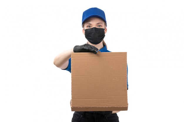 A front view young female courier in blue uniform black gloves and black mask holding food delivery package