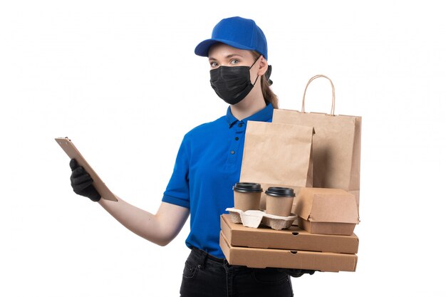 A front view young female courier in blue uniform black gloves and black mask holding food delivery packages and notepad