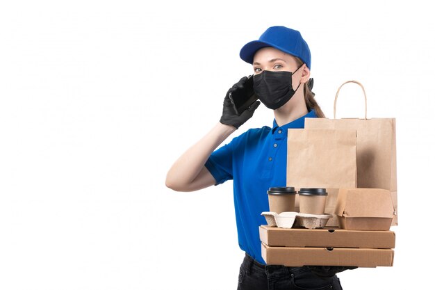 A front view young female courier in blue uniform black gloves and black mask holding food delivery packages and smartphone