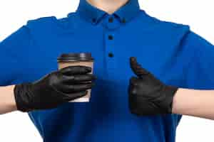 Free photo a front view young female courier in blue uniform black mask and gloves holding coffee cup