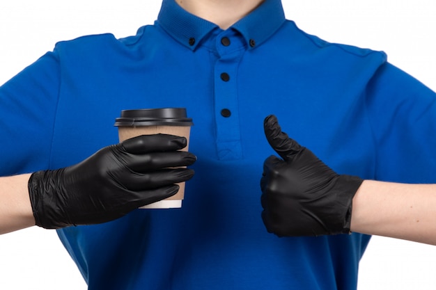 A front view young female courier in blue uniform black mask and gloves holding coffee cup