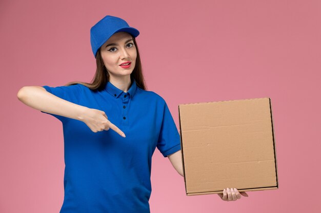 Front view young female courier in blue uniform and cape holding food box and opening it on the pink wall 