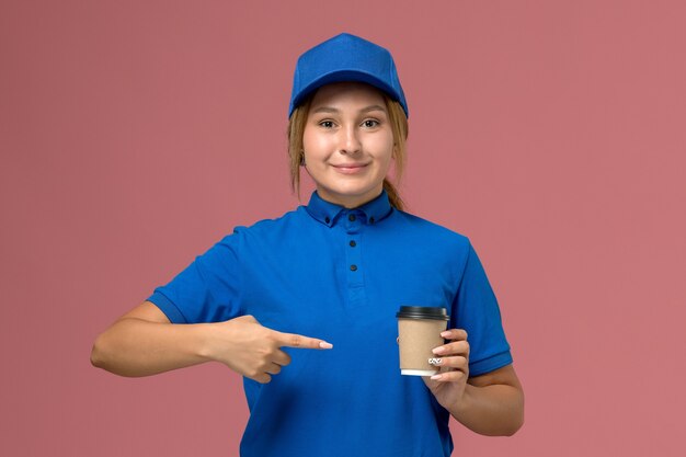 Front view young female courier in blue uniform posing and holding delivery cup of coffee on the pink wall, service uniform delivery woman job