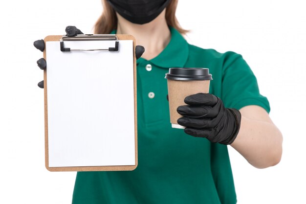 Free photo a front view young female courier in green uniform black gloves and black mask holding coffee cup and notepad