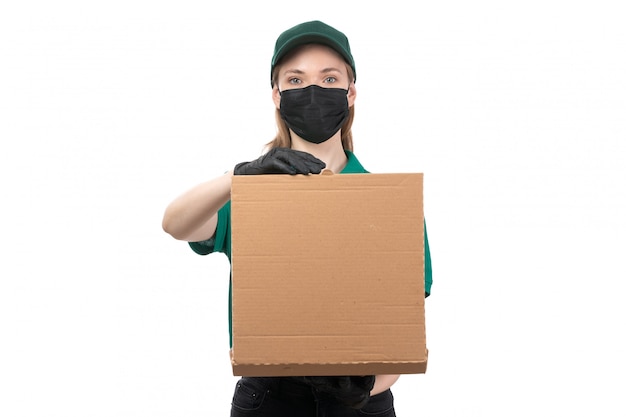 A front view young female courier in green uniform black gloves and black mask holding food delivery package delivering