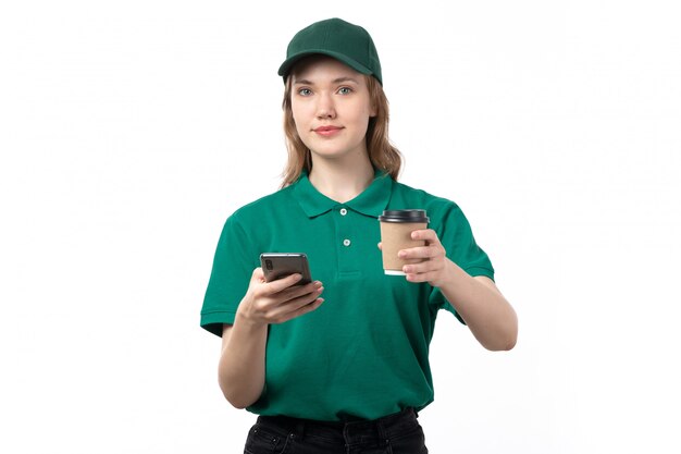 A front view young female courier in green uniform holding coffee cup and using a phone on white