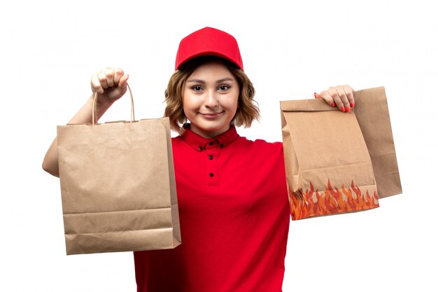 A front view young female courier in red shirt red cap holding delivery packages smiling on the white background uniform service delivering