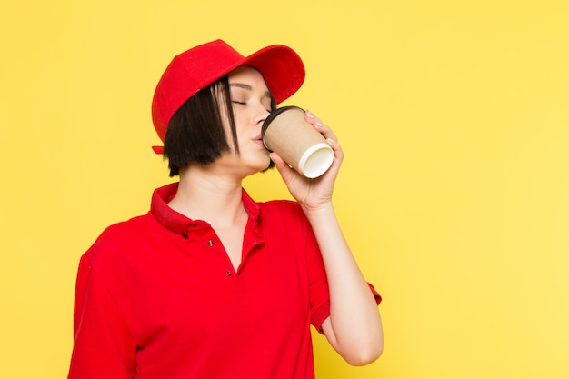 A front view young female courier in red uniform black gloves and red cap drinking coffee