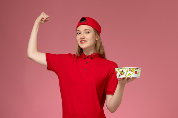 Front view young female courier in red uniform and cape holding delivery bowl flexing on light pink wall