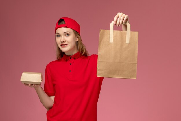 Front view young female courier in red uniform and cape holding delivery food packages on the pink wall