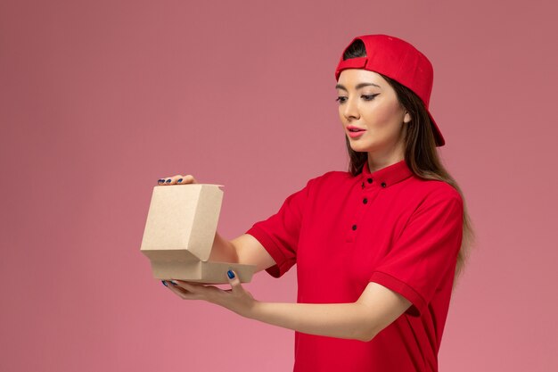 Front view young female courier in red uniform and cape with little delivery food package on her hands on light pink wall