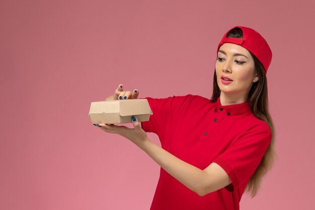 Front view young female courier in red uniform and cape with little delivery food package on her hands on the pink wall