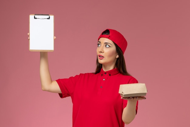 Free photo front view young female courier in red uniform cape with little delivery food package and notepad on her hands on pink wall