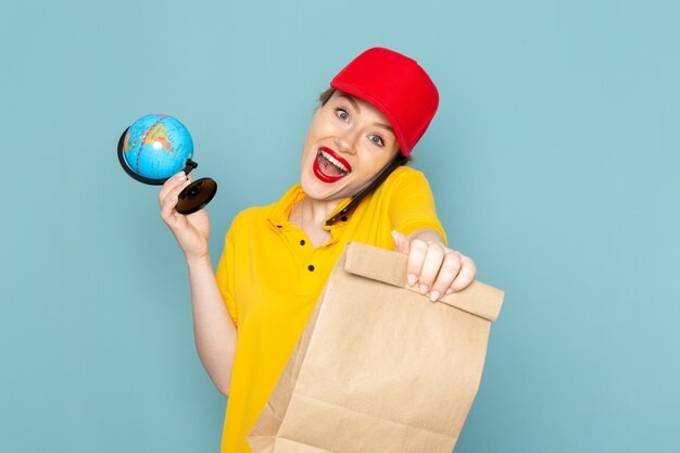 Front view young female courier in yellow shirt and red cape holding globe package talking on the phone on the blue space   