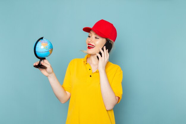 Front view young female courier in yellow shirt and red cape holding little globe talking on the phone on the blue space  