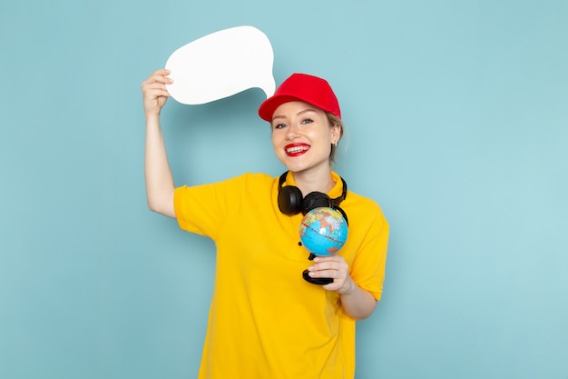 Front view young female courier in yellow shirt and red cape holding little globe and white sign on the blue space   