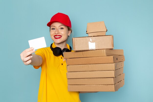 Front view young female courier in yellow shirt and red cape holding multiply  packages and card on the blue space   job