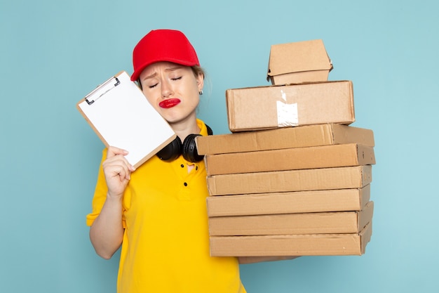 Front view young female courier in yellow shirt and red cape holding multiply  packages and notepad on the blue space   work