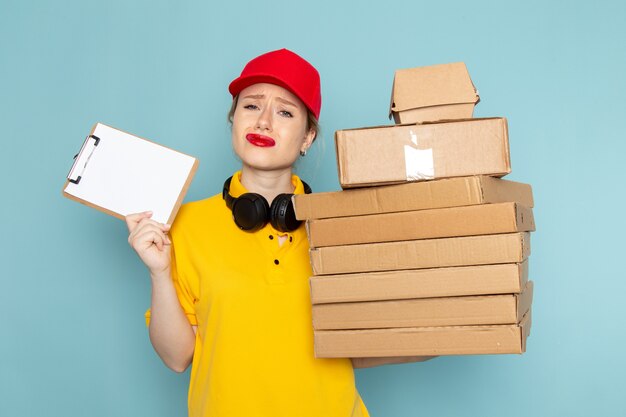 Front view young female courier in yellow shirt and red cape holding multiply  packages and notepad on the blue space  