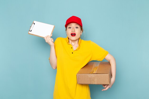 Front view young female courier in yellow shirt and red cape holding  package and notepad on the blue space  job  worker