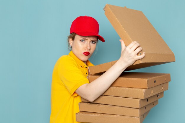 Front view young female courier in yellow shirt and red cape holding  packages opening them on the blue space  