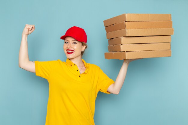 Front view young female courier in yellow shirt and red cape holding  packages smiling on the blue space  worker 