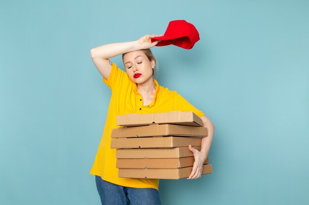 Front view young female courier in yellow shirt and red cape holding  packages taking off cape on the blue space  job 