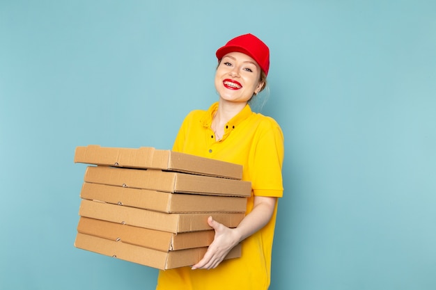 Front view young female courier in yellow shirt and red cape holding  packages with smile on the blue space  job 