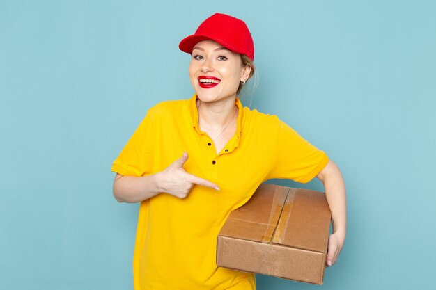 Front view young female courier in yellow shirt and red cape smiling and holding package on the blue space  job 