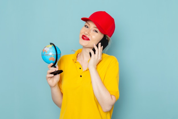 Front view young female courier in yellow shirt and red cape talking on the phone smiling on the blue space  