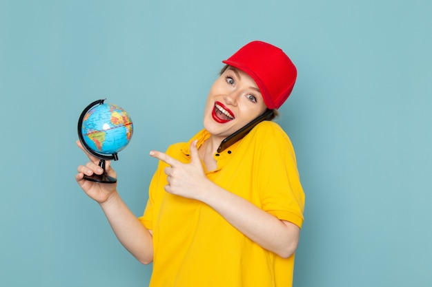 Front view young female courier in yellow shirt and red cape talking on the phone smiling on the blue space  