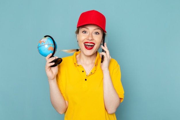 Front view young female courier in yellow shirt and red cape talking on the phone smiling on the blue space  