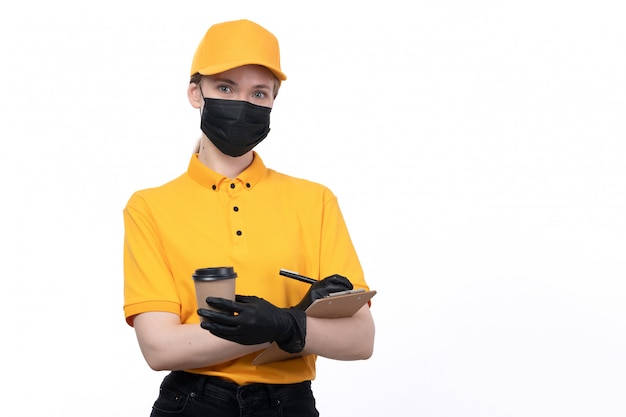 A front view young female courier in yellow uniform black gloves and black mask holding coffee cup and writing order