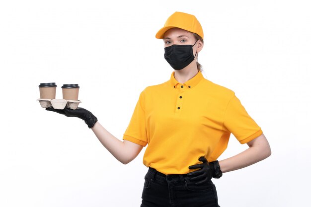 A front view young female courier in yellow uniform black gloves and black mask holding coffee cups delivering