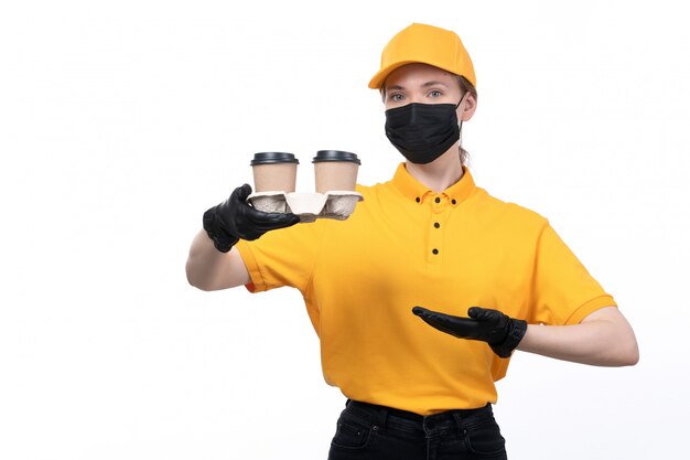 A front view young female courier in yellow uniform black gloves and black mask holding coffee cups