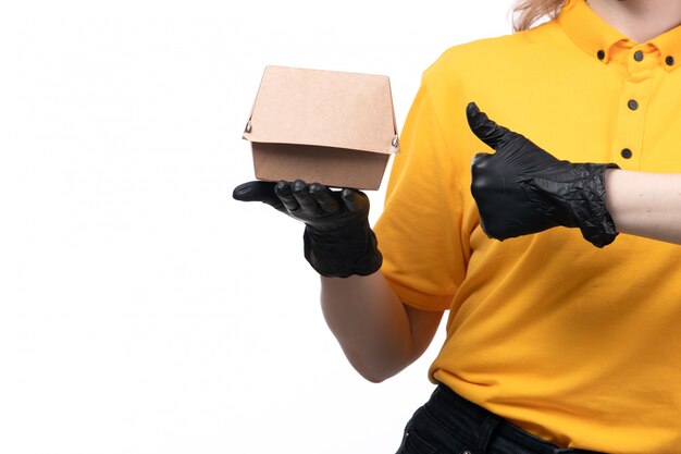 A front view young female courier in yellow uniform black gloves and black mask holding food package