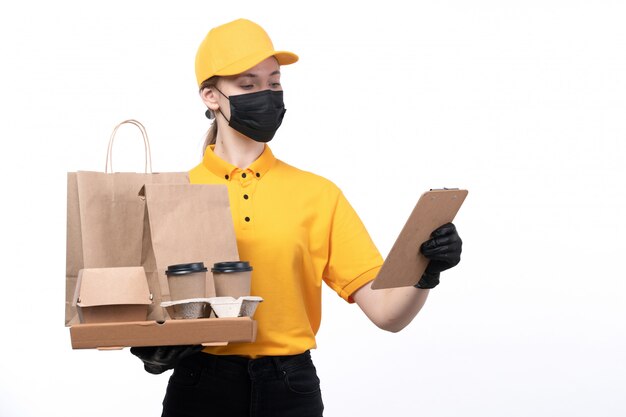 A front view young female courier in yellow uniform black gloves and black mask holding food packages and coffee cups