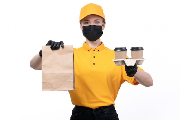 A front view young female courier in yellow uniform black gloves and black mask holding food packages and coffee cups